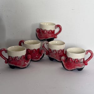 May include: Five ceramic mugs in the shape of red boots with white tops and handles. The mugs have a textured finish and are arranged on a white surface. The mugs are likely for serving hot beverages.