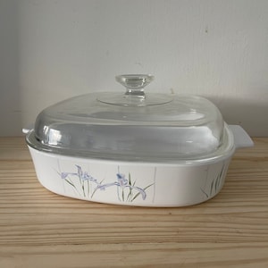 May include: White oval casserole dish with a clear glass lid. The dish features a floral design of purple irises and green leaves. The dish is sitting on a wooden surface.
