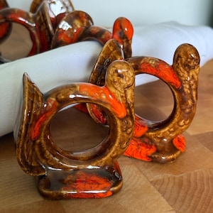 May include: Set of ceramic napkin rings with a turkey design. The rings feature a brown and orange glaze, with a circular opening for the napkin. The turkey-shaped rings are displayed on a wooden surface.