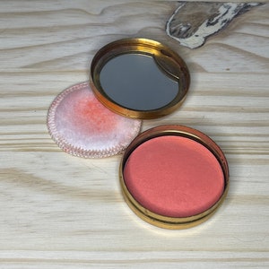 May include: A vintage compact with a gold-tone metal case, open to show a mirror and blush powder. A separate, round, pink powder puff sits next to the compact. The blush is a soft coral colour.