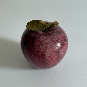 May include: A decorative apple sculpture with a deep red and purple marbled finish. The apple features a brass leaf accent on top. This unique piece is a charming addition to any home decor. (Approx. 8 cm)