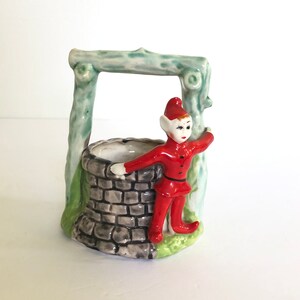 May include: A ceramic figurine of an elf standing next to a well. The elf is wearing a red hat and a red and green outfit. The well is made of grey stone and has a green wooden frame.