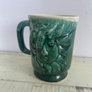 May include: Green ceramic mug with a raised design of a face with leaves. The mug has a white glaze and a handle.