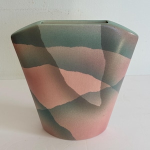 May include: A decorative vase with a geometric design. The vase features a wide, angular shape and is painted with a pattern of overlapping shapes in shades of pink, green, and gray. The top has a rectangular opening.