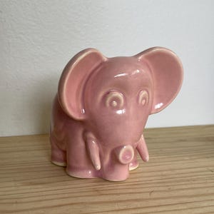 May include: A pink ceramic elephant figurine with white accents around the eyes and trunk. The elephant has a simple, cartoon-like design.