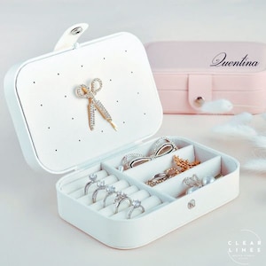 Puede incluir: Caja de joyería blanca con un pequeño lazo en la tapa. La caja tiene varios compartimentos para anillos, pendientes y otras joyas. La caja está abierta y los compartimentos son visibles. La caja está etiquetada con "Quentina" en el lateral.