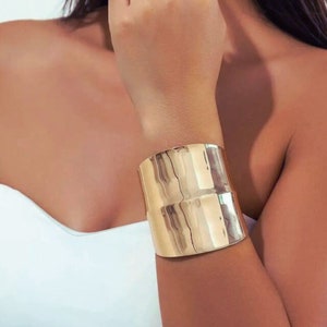 Può includere: Un bracciale a polsino largo color oro. Il bracciale ha una forma rettangolare con una superficie leggermente strutturata. Il gioiello è indossato al polso e riflette la luce, creando un aspetto lucido. Il design generale è audace e moderno.