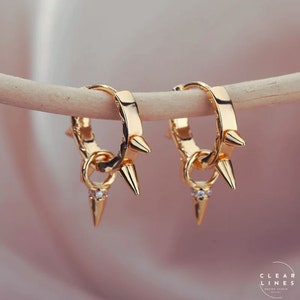 May include: Gold hoop earrings with a small, dangling spike and a single clear stone. The hoops are adorned with multiple spikes.