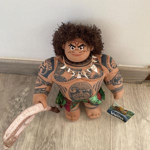 May include: A brown plush toy of Maui from the Disney movie Moana. He is wearing a grass skirt and a necklace with a shell pendant. He has a large tattoo on his chest and is holding a wooden club. The toy has a tag that says "Moana".