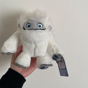 Puede incluir: Peluche blanca de Yeti con ojos azules y boca gris. El juguete sostiene una etiqueta que dice "The Snowman" y tiene una marca CE.