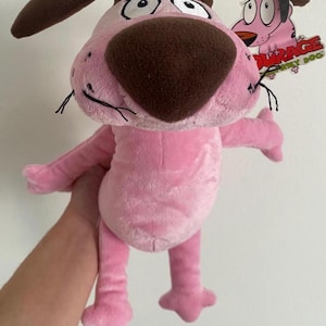 Puede incluir: Juguete de peluche rosa Courage el perro cobarde con orejas marrones y una nariz marrón. El juguete tiene una etiqueta que dice "Courage el perro cobarde".