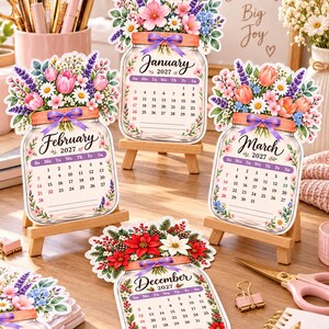 May include: Four die-cut mason jar calendars for January, February, March, and December 2027. Each features a floral arrangement, a purple ribbon, and a monthly grid. The calendars are displayed on small wooden easels on a desk.