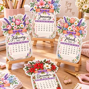 May include: Four mason jar-shaped 2027 calendars displayed on small wooden easels. Each calendar features a unique floral arrangement at the top, a purple ribbon tied around the jar neck, and a monthly grid for January, February, March, and December.