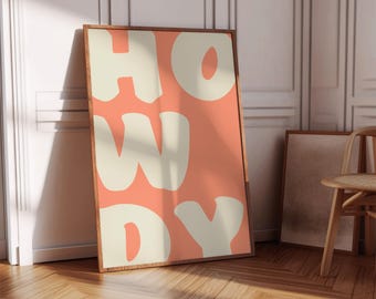 May include: A framed art print with the word "HOWDY" in large, cream-colored letters against a peach background. The wooden frame complements the warm tones of the artwork. The print leans against a white wall and wooden floor.