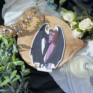 Może przedstawiać: Naklejka przedstawiająca parę z czarnymi skrzydłami, kobieta w różowej sukience, a mężczyzna w czarnej koszuli i spodniach. Naklejka znajduje się na drewnianej powierzchni.