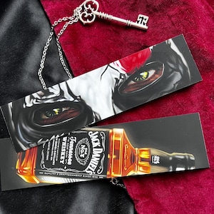Puede incluir: Dos marcapáginas ilustrados y un colgante de llave plateada. Uno muestra una botella de whisky Jack Daniel's. El otro presenta una ilustración de un rostro con ojos verdes. La llave está en una cadena.