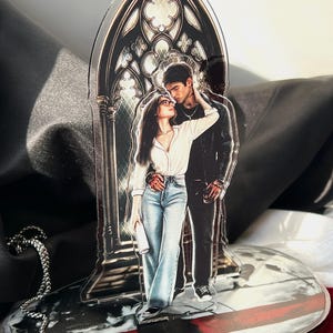 May include: A clear acrylic standee featuring a printed illustration of two figures standing before a gothic arched window. One figure wears a white shirt and blue jeans, while the other wears a black jacket and dark trousers. The base is oval.