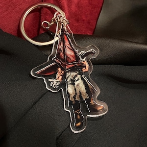 May include: A clear acrylic keychain featuring a character with a red pyramid-shaped helmet and a large knife. The character is depicted in a detailed, artistic style, with brown boots and a long coat. The keychain has a silver ring.