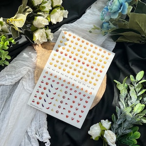 May include: A sheet of white sticker paper with a variety of colorful emojis. The emojis include smiling faces, hearts, fire, lips, cherries, peaches, bananas, eggplants, and peppers.
