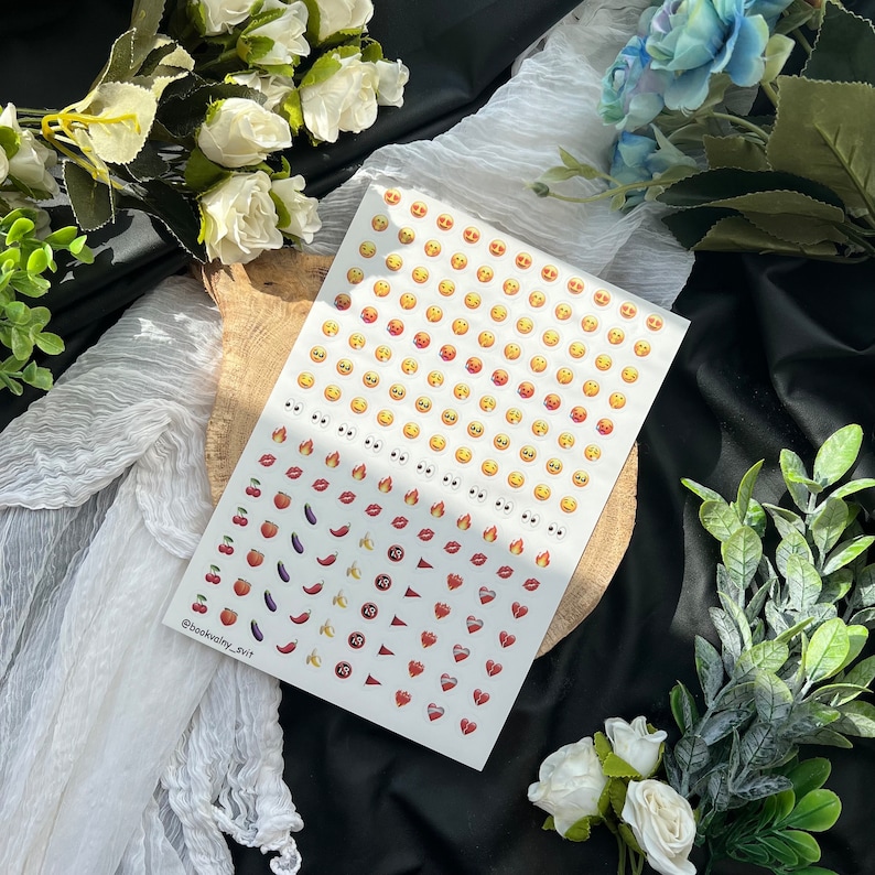 May include: A sheet of white sticker paper with a variety of colorful emojis. The emojis include smiling faces, hearts, fire, lips, cherries, peaches, bananas, eggplants, and peppers.