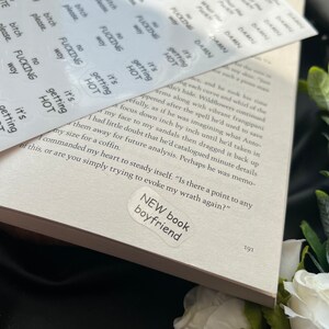 May include: A close-up of a book with a sticker that says "NEW book boyfriend" on the cover. The sticker is white with black text. The book is open to a page with text about a character's wrath.