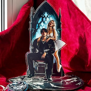 May include: An acrylic standee featuring a dark-haired person in a black jacket sitting on a chair in front of a gothic archway, with a blonde person in a white dress standing behind them. A small, separate acrylic charm with a graphic design is included.
