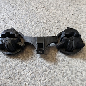 Op de afbeelding: Een zwart, 3D-geprint object met twee sierlijke, schedelachtige vormen die door een centrale balk zijn verbonden. Het object heeft een textuuroppervlak en lijkt van plastic te zijn. Het ontwerp is symmetrisch en gedetailleerd.