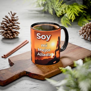 Puede incluir: Taza de café con asa negra con el texto "Soy mi Mejor Aliada" en blanco y naranja. La taza presenta una silueta de una persona de pie sobre una montaña con un fondo de puesta de sol. La taza está sobre una tabla de cortar de madera.
