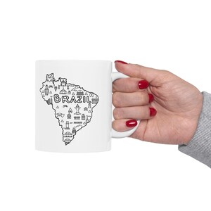 May include: A white ceramic mug featuring a black line art map of Brazil. The map is filled with various small, detailed illustrations of landmarks and the word BRAZIL printed in bold, capital letters across the center of the country shape.