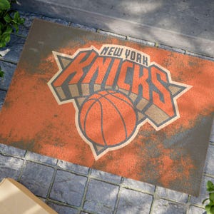 Può includere: Uno zerbino rettangolare caratterizzato da una superficie testurizzata color arancione e grigio dall'aspetto usurato, con il logo dei New York Knicks stampato al centro. Il design include il nome della squadra in grassetto sopra un pallone.