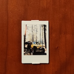 May include: A white polaroid photo of a yellow Jeep in a forest setting, attached to a brown wooden surface. The Jeep is parked near trees, with a tent-like structure visible. The photo is held in place by two white clips.