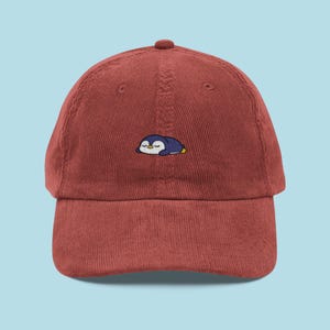 May include: A brick-red corduroy baseball cap with an embroidered sleeping penguin design. The penguin is navy blue with a white belly and yellow details. The cap has a curved peak and a button on top.
