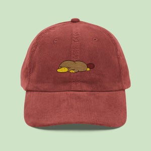 May include: A maroon corduroy baseball cap with an embroidered cartoon platypus in brown, yellow, and red. The platypus is lying down. The cap has a curved peak and an adjustable strap.