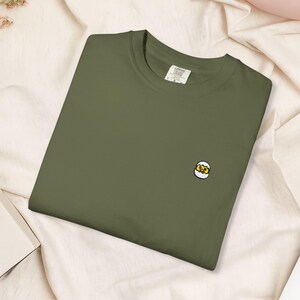 May include: Olive green t-shirt with a small embroidered egg design on the front. The shirt is made of a soft material and has a classic crew neck. The tag reads "Comfort Colors".