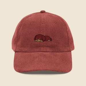 May include: A terracotta corduroy baseball cap with an embroidered dachshund puppy design. The puppy is depicted lying down. The cap has a curved peak and a button on top. The cap is a stylish accessory.