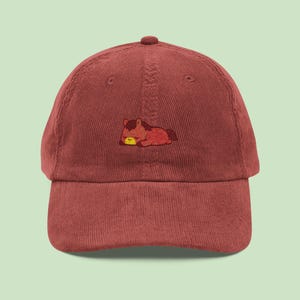 May include: A maroon corduroy baseball cap with an embroidered cartoon horse in brown, red, and yellow. The horse is depicted lying down, creating a playful design on the front. The cap has a curved peak and an adjustable strap for a comfortable fit.
