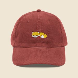 May include: A brick-red corduroy baseball cap with an embroidered sleeping fox. The fox is yellow and white with a red nose. The cap has a curved brim and a button on top.