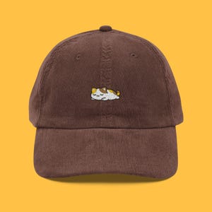 May include: A brown corduroy baseball cap with an embroidered sleeping cat design in white, brown, and yellow. The cap has a curved peak and a button on top. A fashionable accessory for cat enthusiasts.