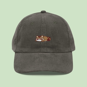 May include: A dark olive green corduroy baseball cap with an embroidered raccoon design. The raccoon is brown, tan, and white, in a lying down pose. The cap has a curved peak and an adjustable strap.