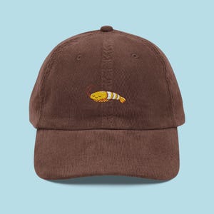 May include: A brown corduroy baseball cap with an embroidered cartoon shrimp. The shrimp is yellow, white, and orange, with closed eyes. The cap has a curved brim and a button on top. The hat is a popular casual accessory.