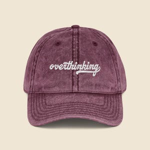 May include: A maroon baseball cap with the word "overthinking" embroidered in white script across the front. The cap has a curved brim and a vintage, worn look. The hat is made of a soft, textured fabric.