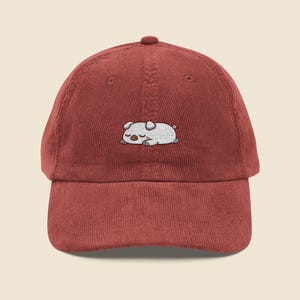 May include: A brick-red corduroy baseball cap with an embroidered sleeping pig design in white and grey. The cap has a curved peak and a top button. A casual accessory for everyday wear.