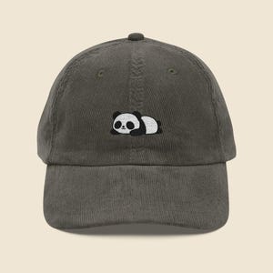 May include: A dark olive green corduroy baseball cap with an embroidered panda bear. The panda is black and white and is lying down. The cap has a curved peak and an adjustable strap at the back.
