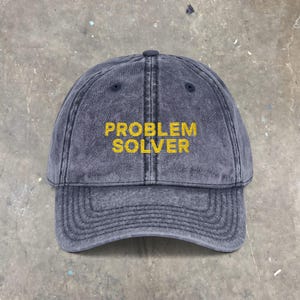 May include: A faded navy blue baseball cap with the embroidered text "PROBLEM SOLVER" in yellow. The cap has a curved brim and a button on top. The hat is made of a soft, worn material.