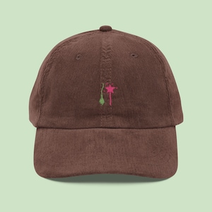 May include: A brown corduroy baseball cap with a small embroidered design of a pink and green flower. The cap has a curved brim and a rounded top.