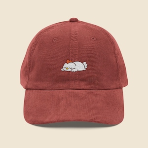 May include: A reddish-brown corduroy baseball cap. It has an embroidered design of a sleeping chicken in white, with a yellow beak and a red comb. The cap has a curved peak and a button on top. The cap is a casual accessory.