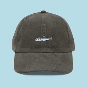 May include: An olive green corduroy baseball cap with a small embroidered fish design. The fish is depicted in blue and white, with closed eyes. The cap is a classic style, suitable for casual wear.