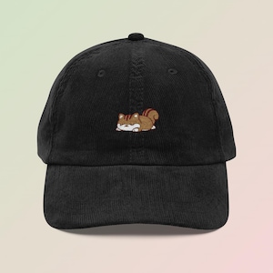 May include: Black corduroy baseball cap with an embroidered cartoon squirrel in brown, tan, and white. The squirrel is lying down. The cap has a curved peak and a button on top.