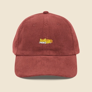 May include: A deep red corduroy baseball cap with a small embroidered design of a sleeping tabby cat. The cat is orange and yellow. The cap has a curved peak and an adjustable strap for a comfortable fit. The hat is made of corduroy.
