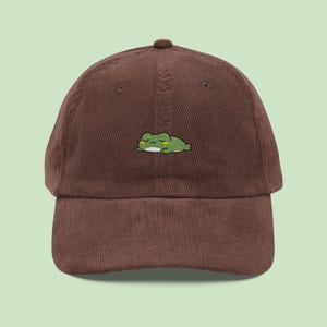 May include: A brown corduroy baseball cap with an embroidered green frog design. The frog is lying down with its eyes closed. The cap has a curved peak and a button on top.